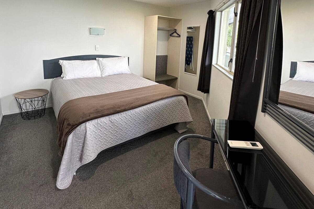 Une chambre d’hôtel avec un lit, un bureau, une chaise, un miroir et une fenêtre avec des rideaux.