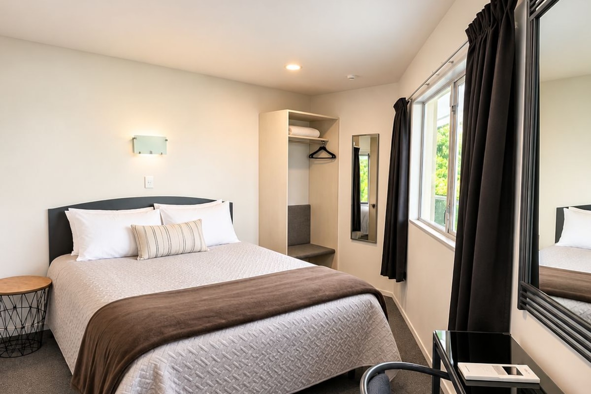 Une chambre d’hôtel avec un lit, un bureau, une chaise, un miroir et une fenêtre avec des rideaux.