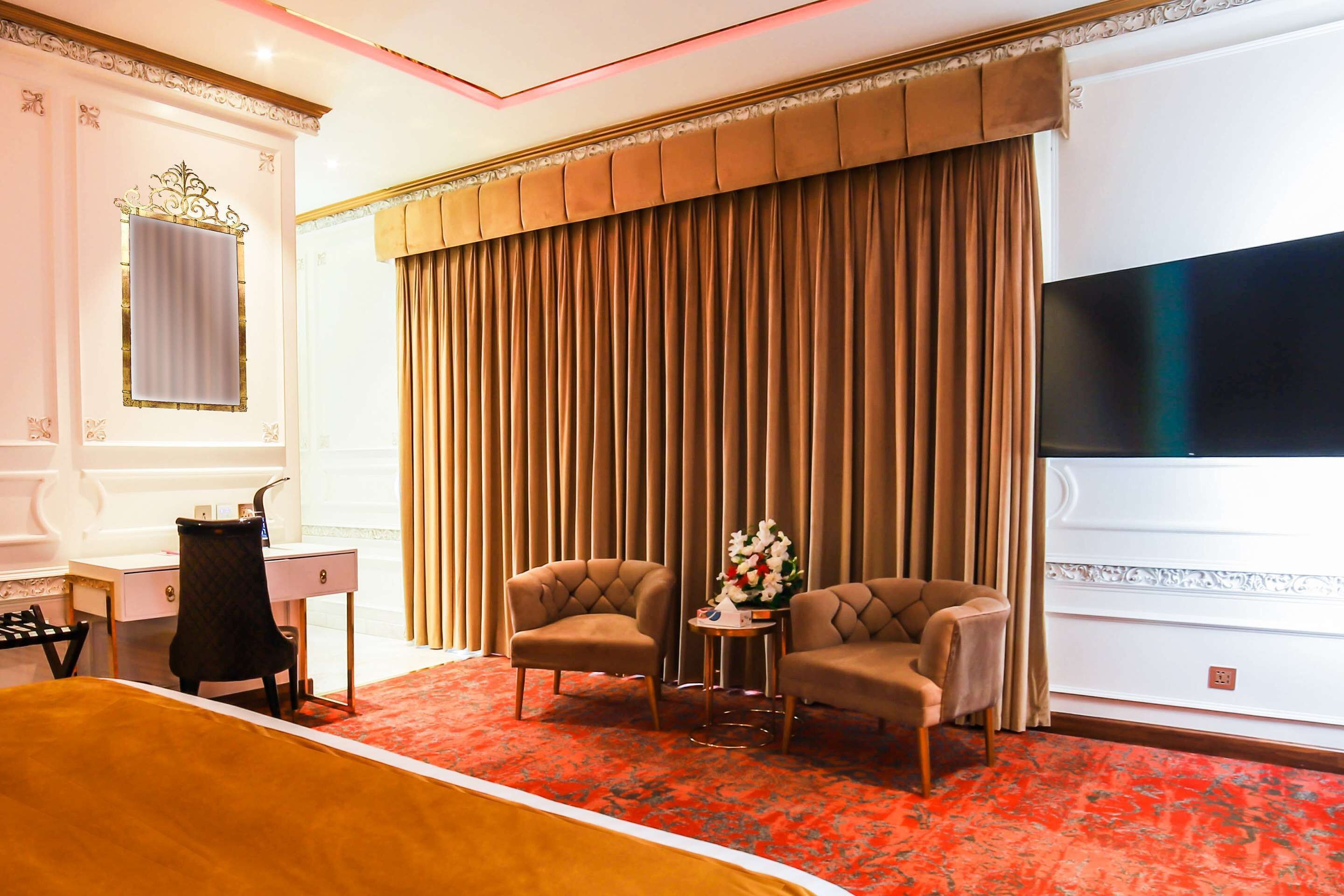 A hotel room with a large bed, a desk with a chair, two armchairs, a small table with a vase of flowers, and a large TV on the wall.