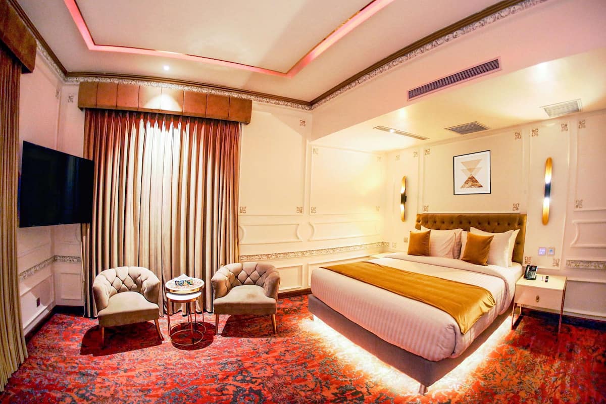 Hotellihuone, jossa on suuri sänky, kaksi nojatuolia, pieni pöytä, televisio ja punainen matto.