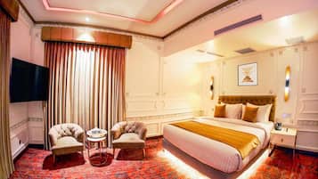 A hotel room with a large bed, two armchairs, a small table, a TV, and a red carpet.