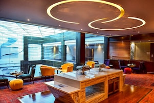 A modern lobby with a marble reception desk, yellow chairs, and large windows offering views of the city.
