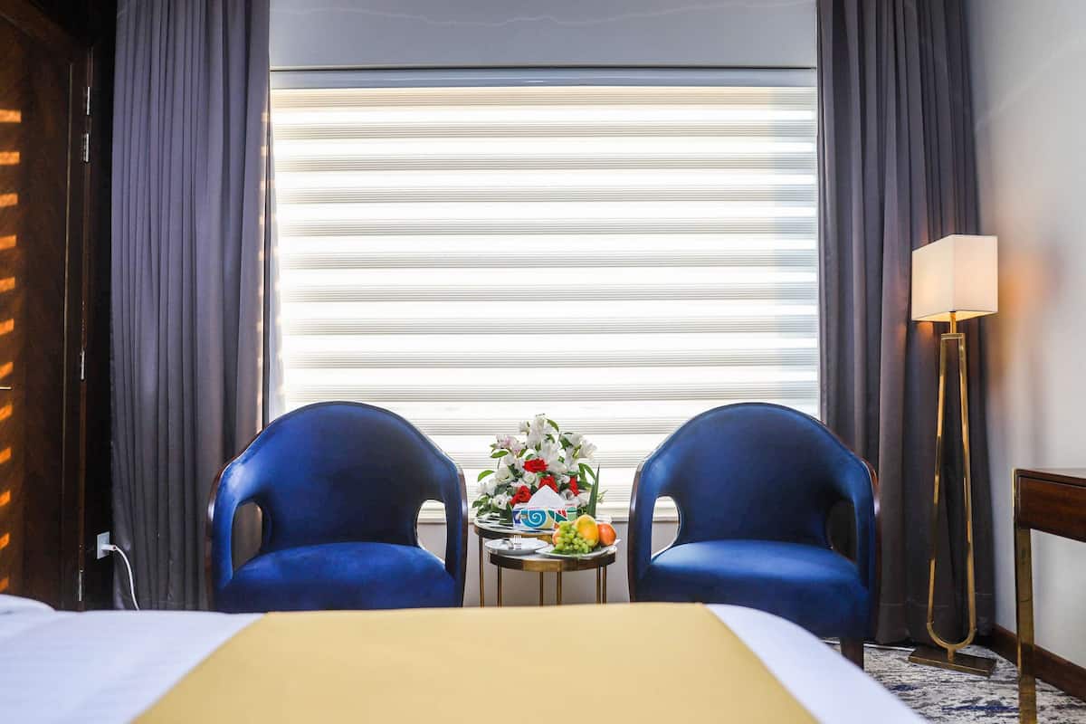 Hotellihuone, jossa on sänky, kaksi sinistä tuolia, pieni pöytä, jossa on hedelmiä ja kukkia, lamppu ja ikkuna, jossa on sälekaihtimet.