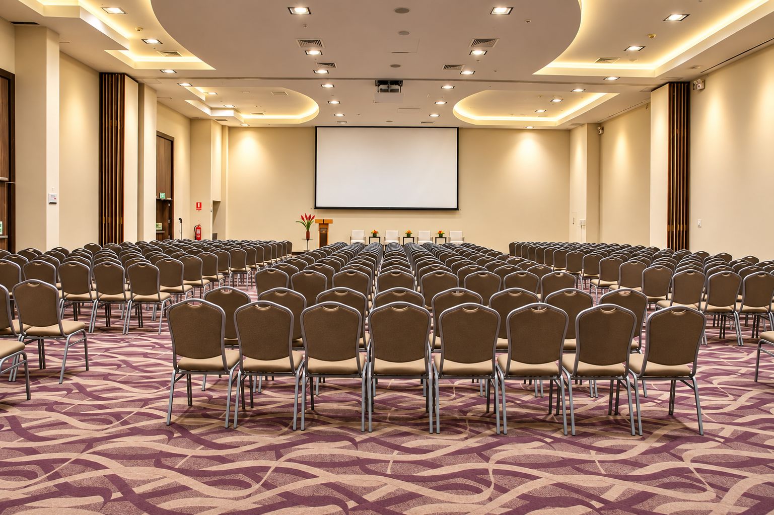 Sala de conferência com fileiras de cadeiras, uma tela de projetor e piso coberto de carpete.