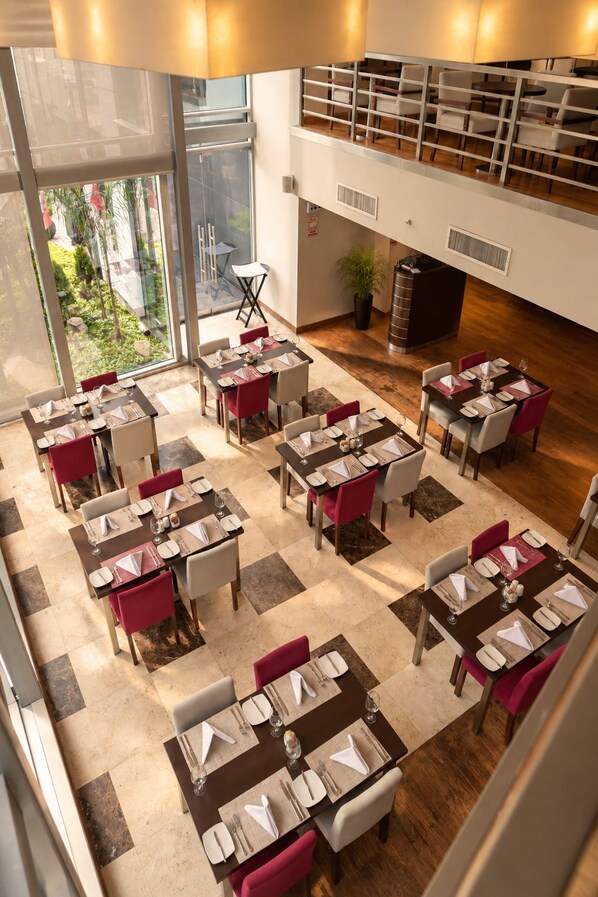 Zona pranzo moderna con diversi tavoli e sedie, vista sul verde esterno e un soppalco con posti a sedere.