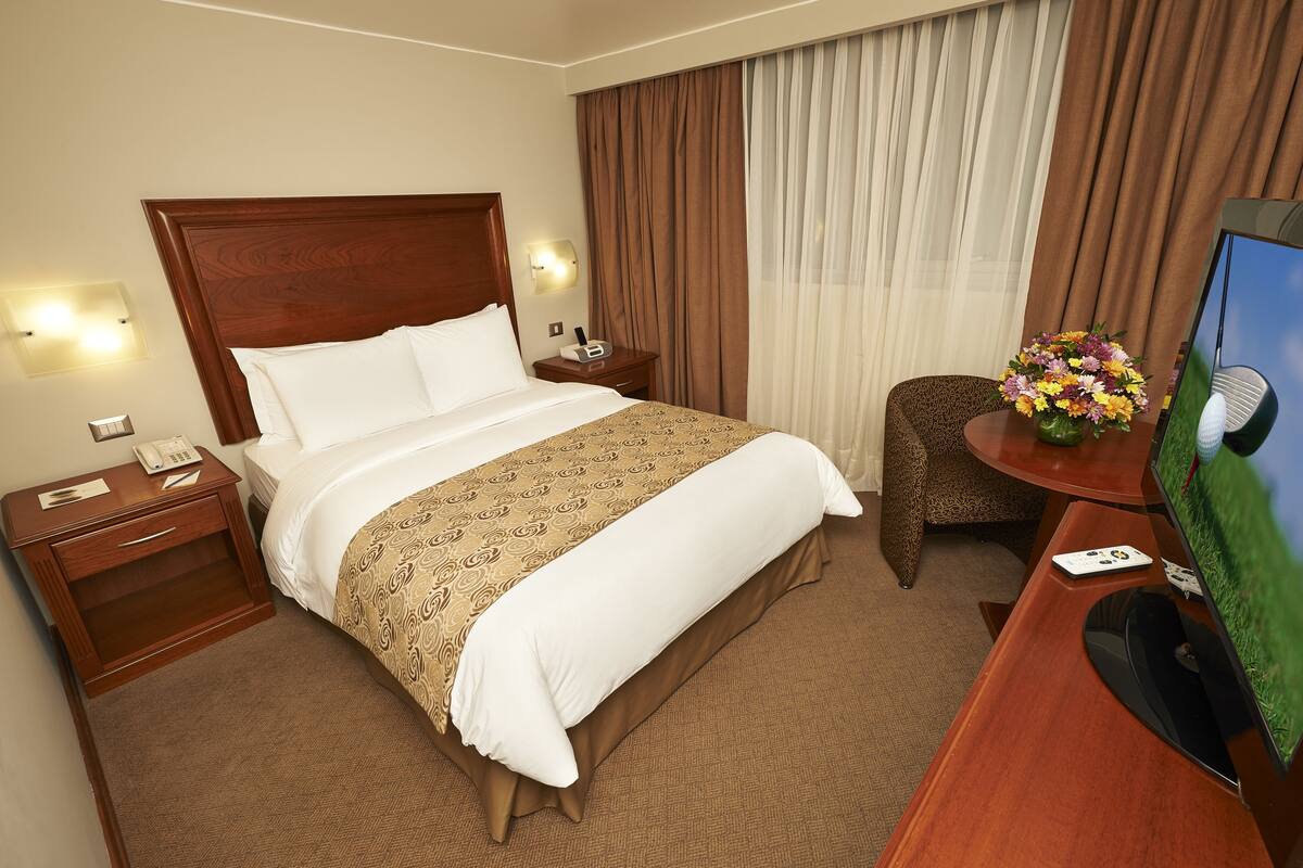 Habitación de hotel con una cama grande, una mesita de noche de madera, un escritorio con televisor y una silla.