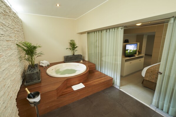 A modern bathroom with a wooden bathtub, a stone wall, a potted plant, a sink, and a television. - Sol de Oro Hotel & Suites (Lima)