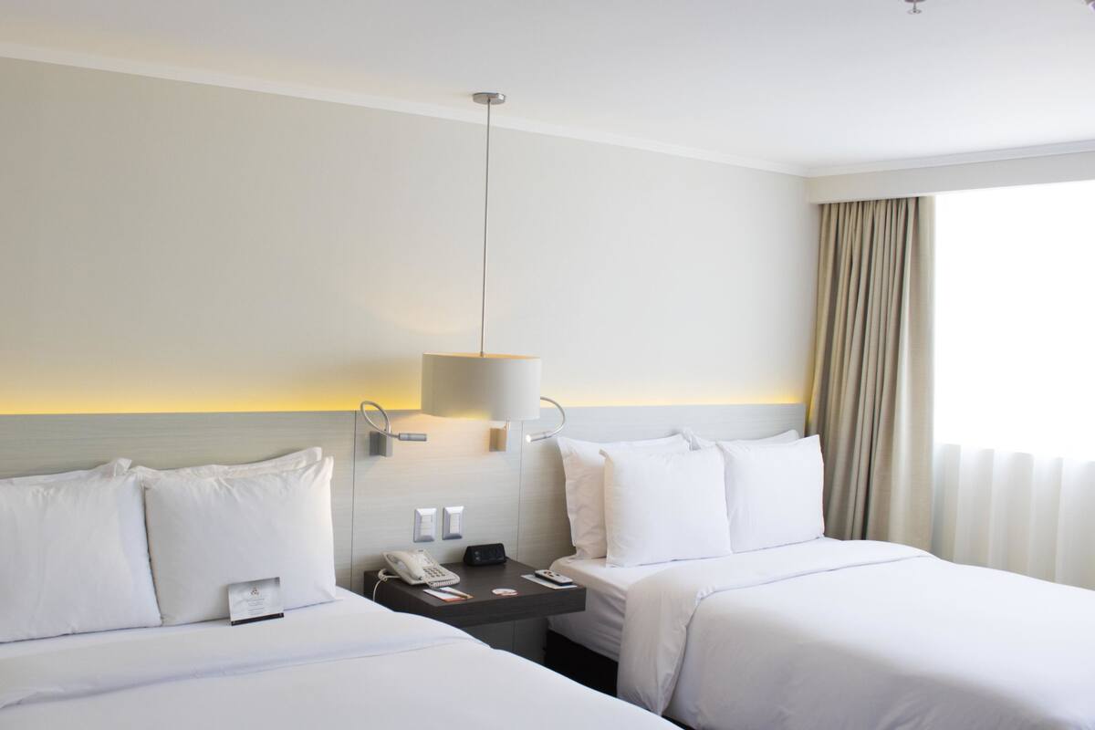 Habitación de hotel con dos camas, una mesita de noche con un teléfono y una lámpara, y vista al mar por la ventana.