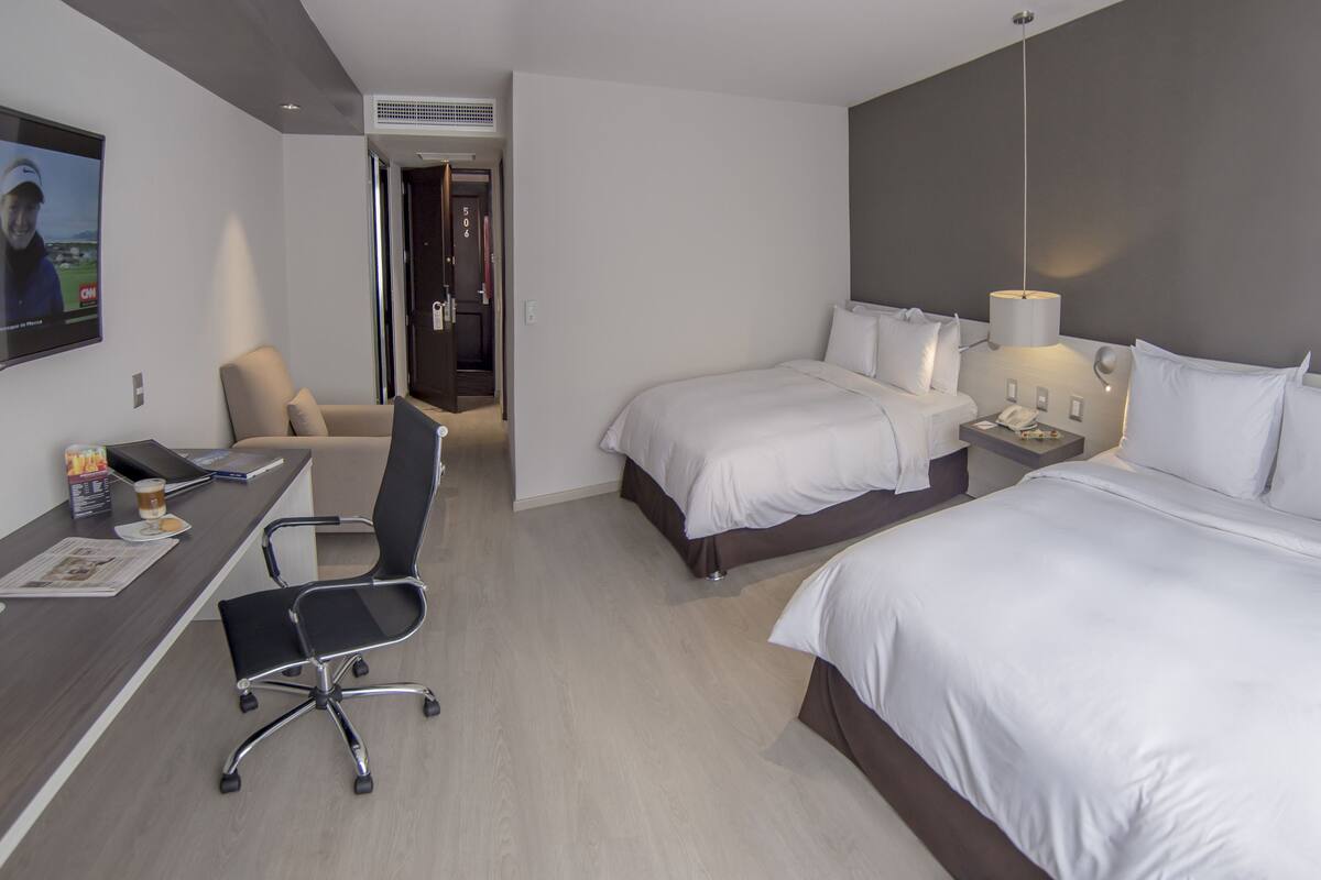 Habitación de hotel con dos camas, un escritorio con silla, un televisor y una lámpara.