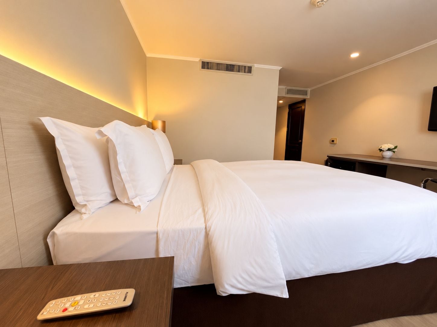 A hotel room with a large bed, a bedside table with a remote control, a wall-mounted TV, and a small vase with flowers on the dresser.