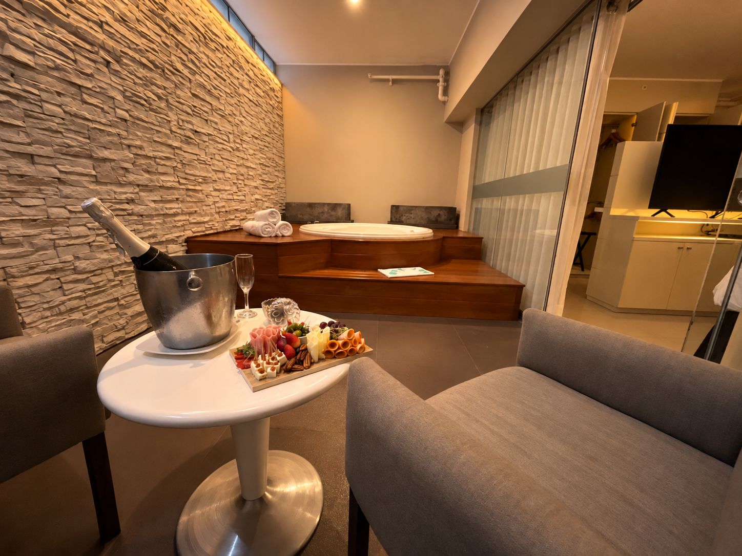 A modern hotel room with a stone wall, a wooden seating area, a small table with a bottle of champagne, glasses, and a charcuterie board.