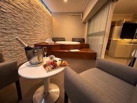 A modern hotel room with a stone wall, a wooden seating area, a small table with a bottle of champagne, glasses, and a charcuterie board.