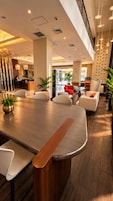 A modern hotel lobby with a long conference table, seating area with chairs, and a reception desk.
