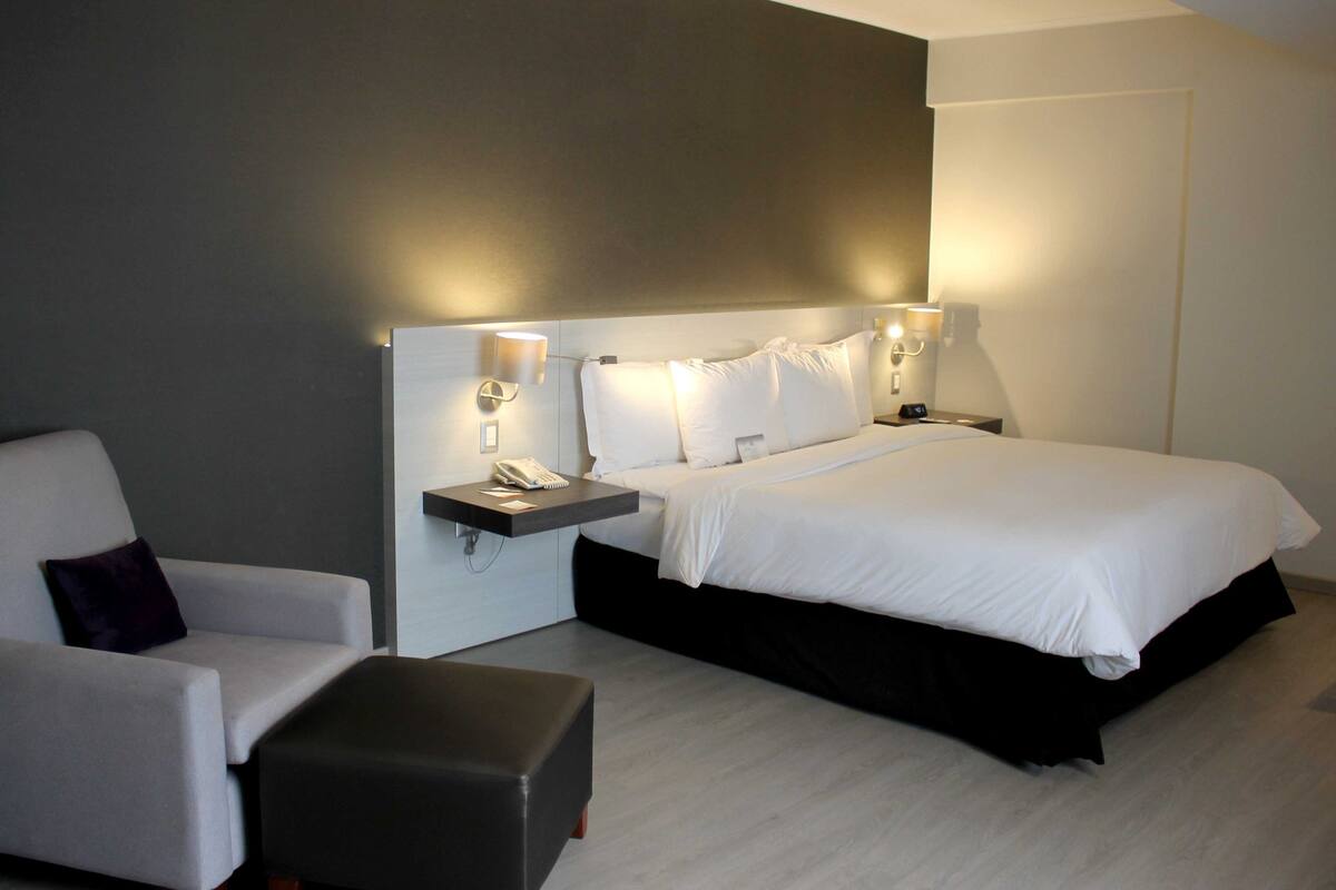 Una habitación de hotel moderna con una cama, una mesita de noche con lámpara, una silla y un pequeño otoman.