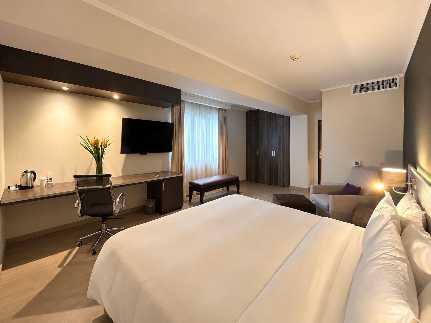 Ett hotellrum med en stor säng, ett skrivbord med en platt-TV, en sittgrupp med en bänk och en garderob.