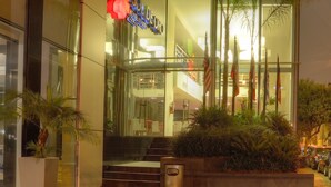 Un edificio moderno con ingresso in vetro, una insegna con la scritta "Hotel de Oro" e un fioriere con piante e bandiere all'esterno.