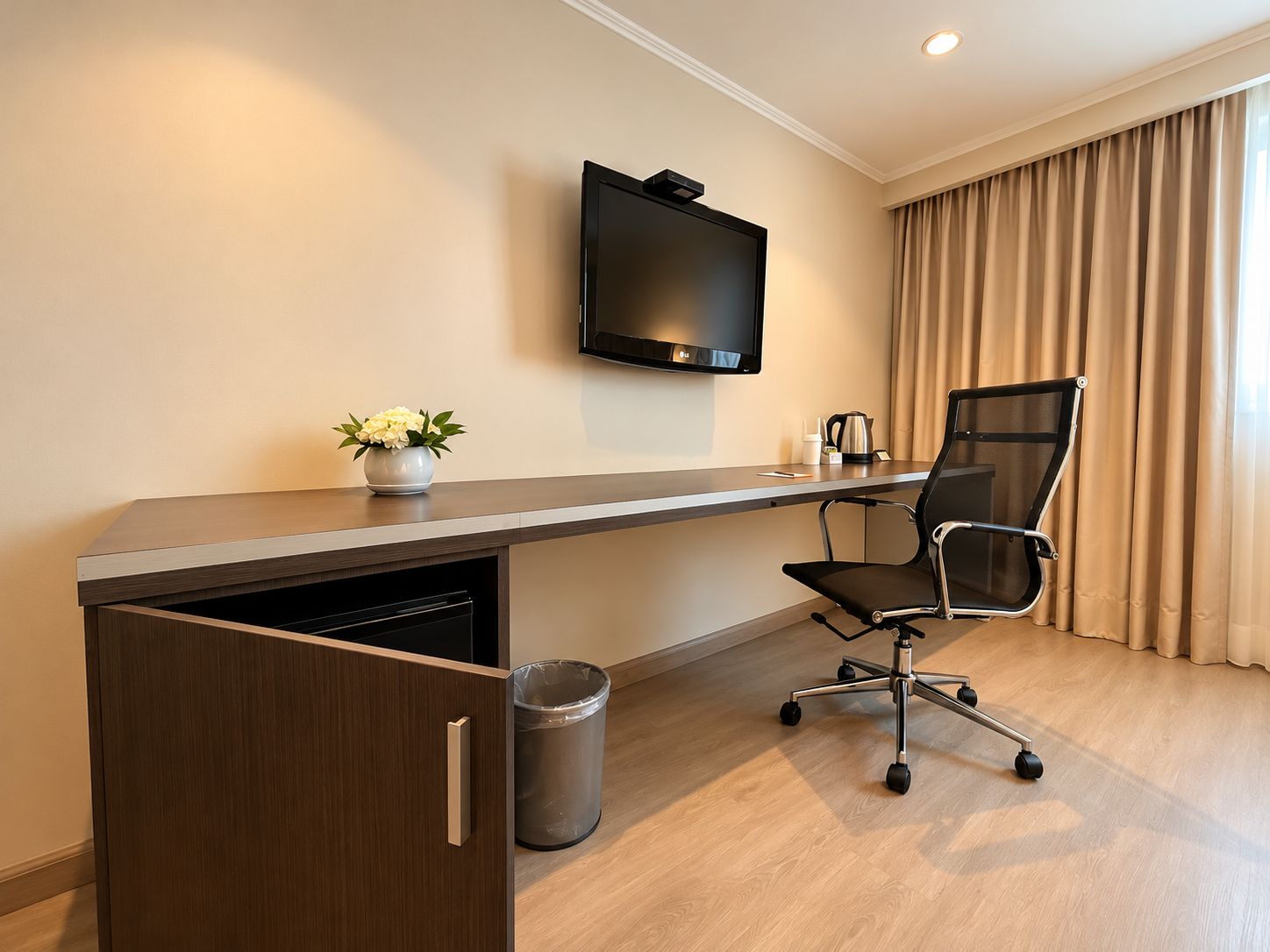 Habitación de hotel con un escritorio de madera, una silla de oficina negra, un televisor montado en la pared, una pequeña planta en maceta y un cubo de basura.