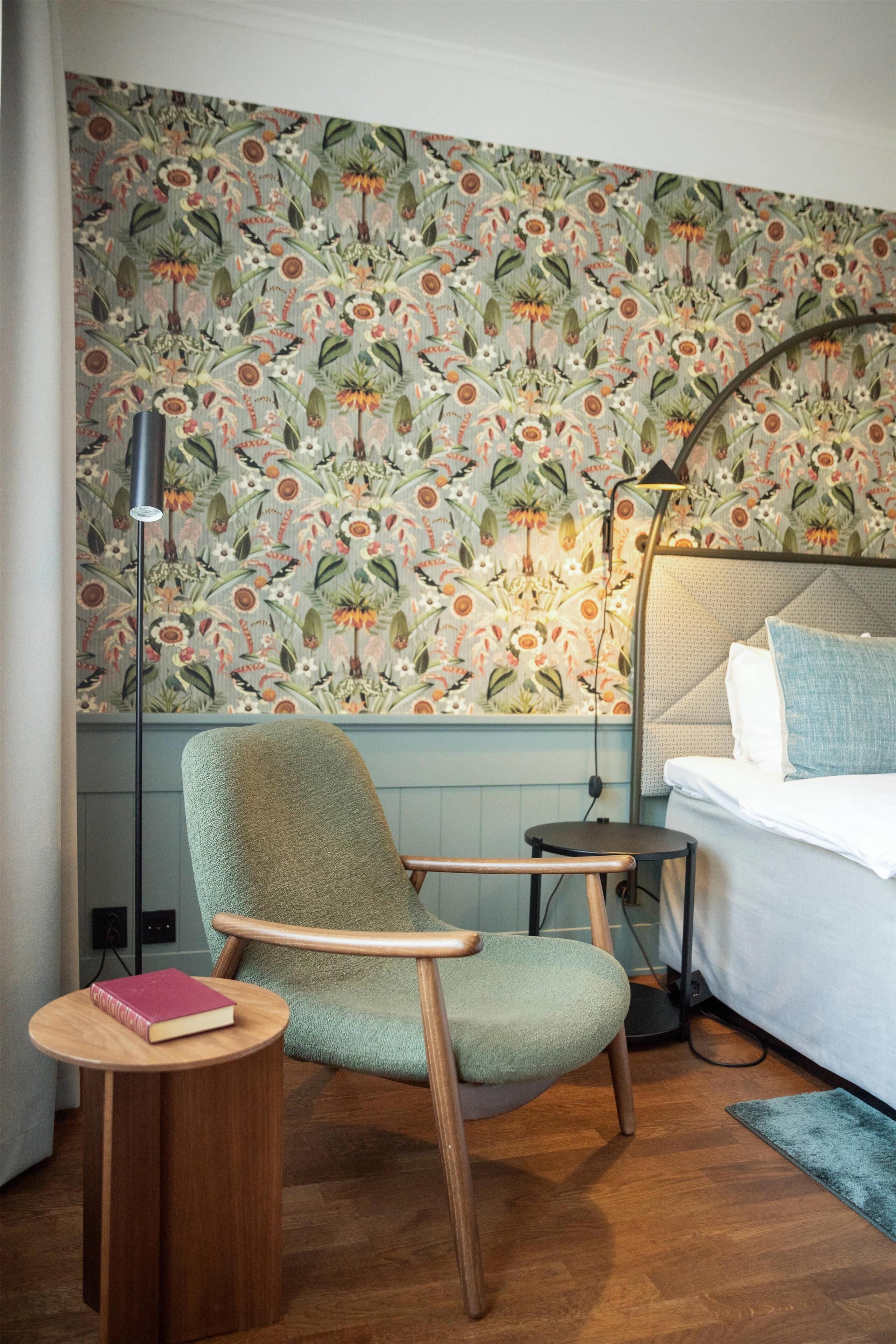 A room with a floral wallpaper, a green armchair, a small wooden side table with a book, a bedside table with a lamp, and a bed with a blue pillow.