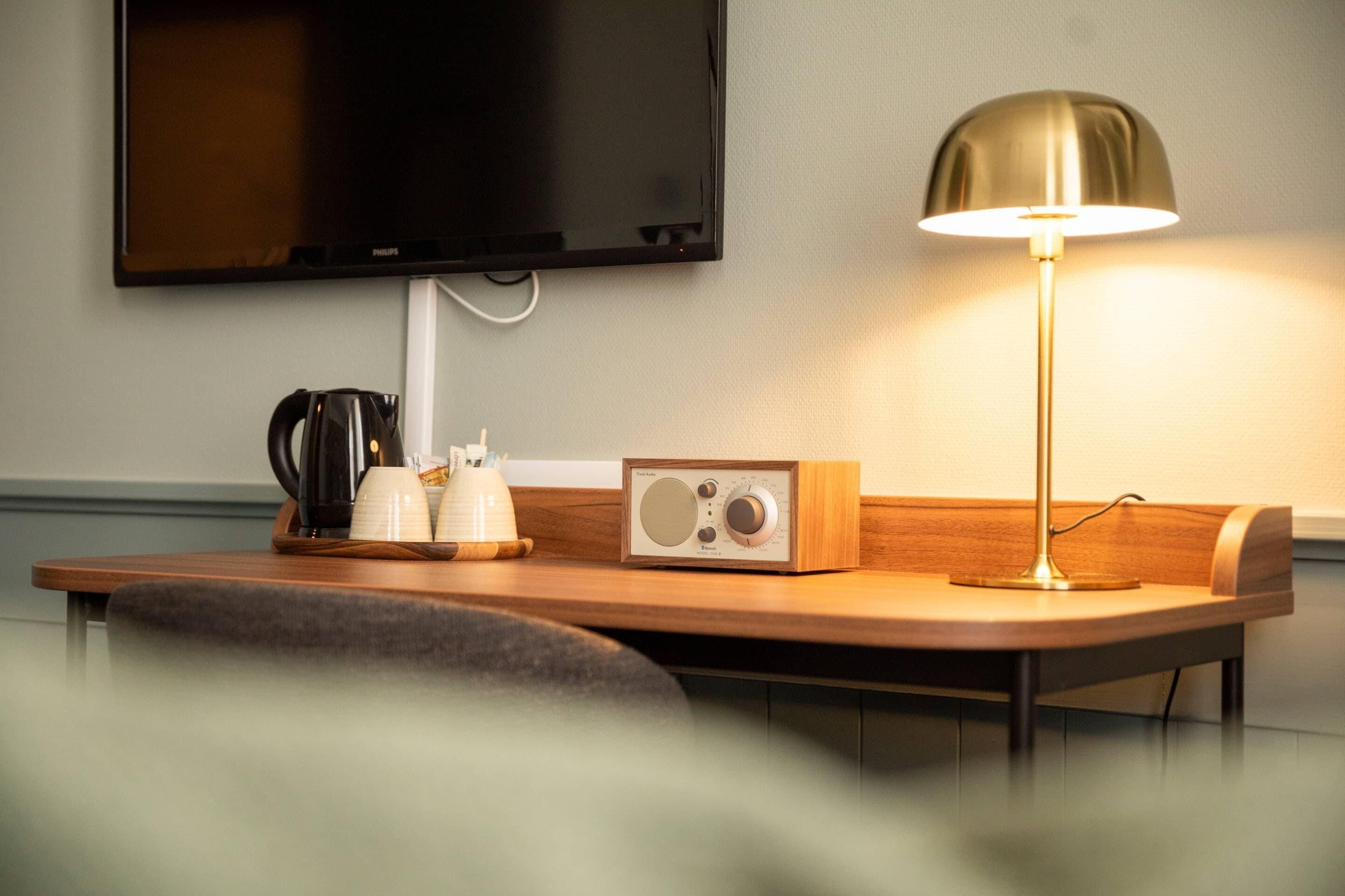 A modern hotel room with a wooden desk, a flat-screen TV mounted on the wall, a radio, and a lamp.