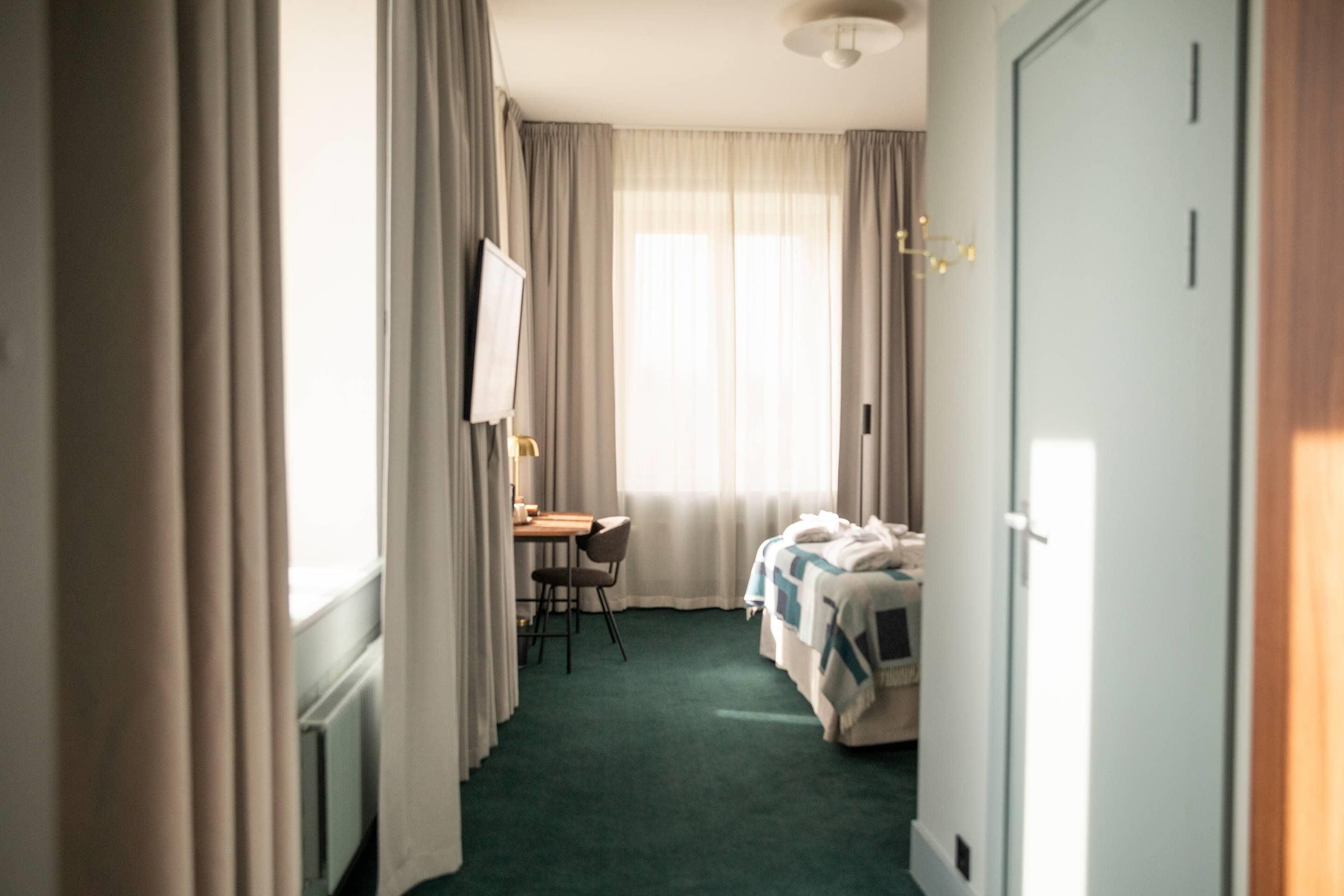A hotel room with a bed, a desk, and a chair. There is a window with curtains and a view of the outside.