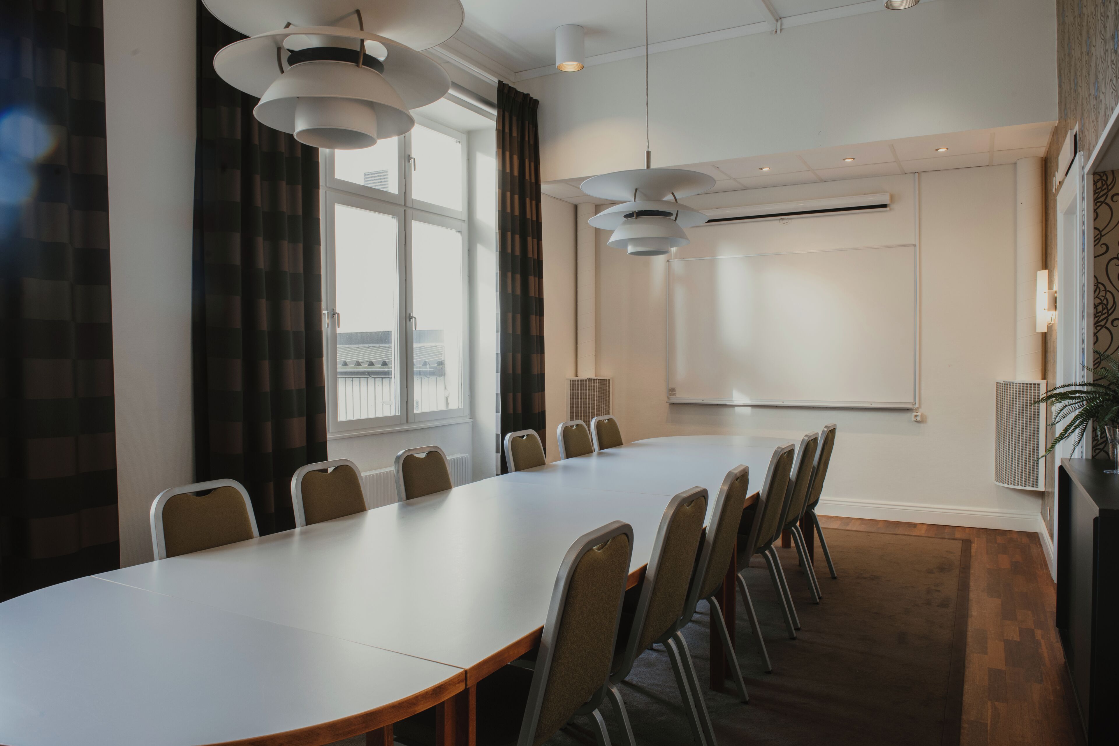 Ett konferensrum med ett långt bord, stolar, en whiteboard och ett stort fönster med gardiner.