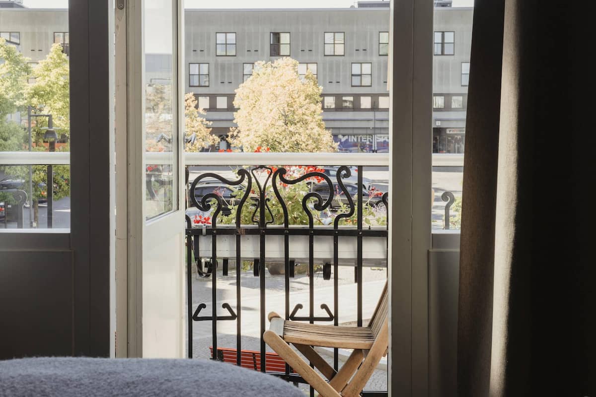 En balkong med en trästol, ett metallräcke och utsikt över byggnader och träd.