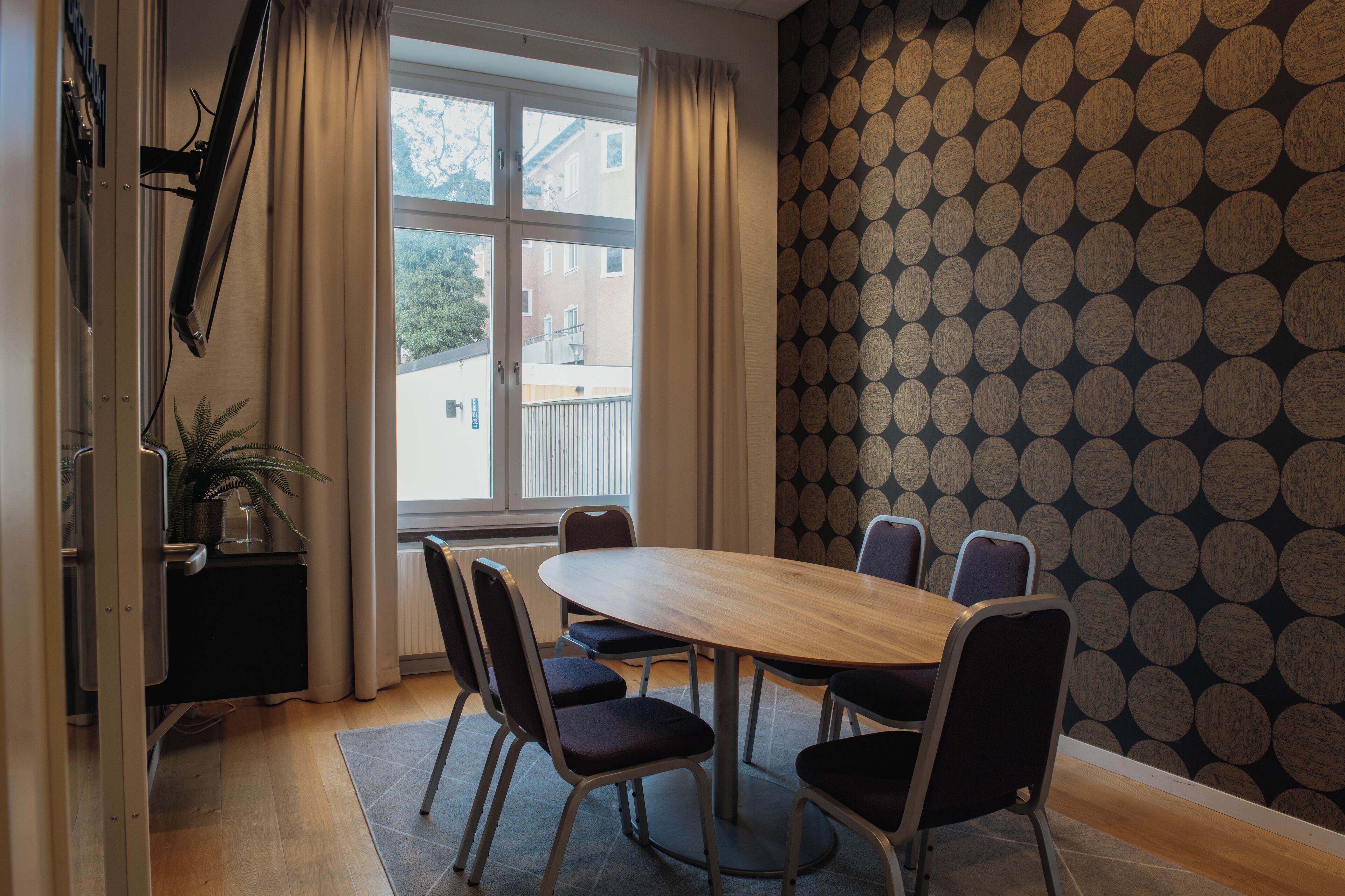 Une salle de conférence avec une table ronde, six chaises, une plante en pot et une fenêtre avec des rideaux.
