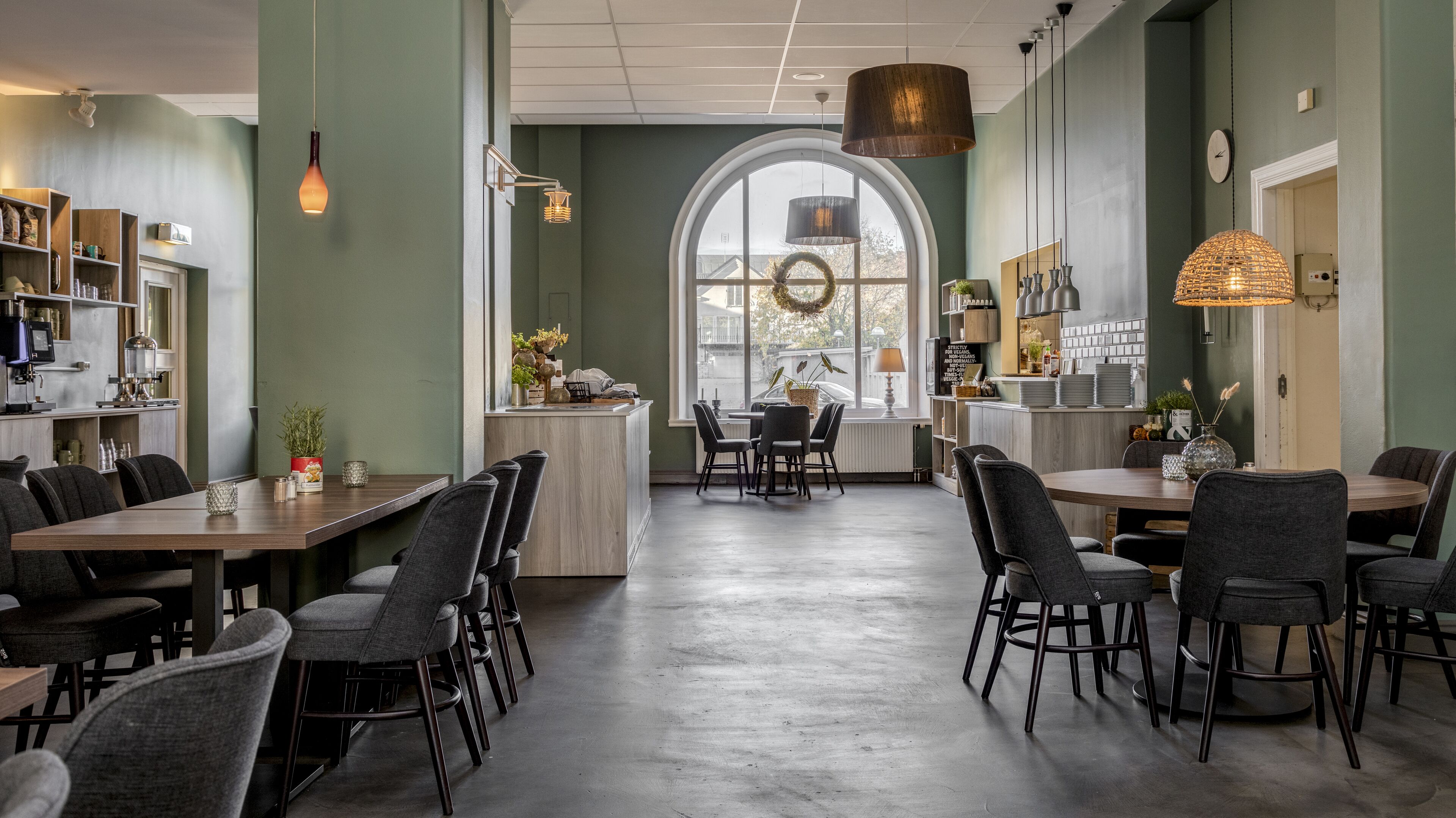 En moderne restaurant med et centralt spiseområde, køkken synligt til venstre, og et stort buet vindue med en krans.