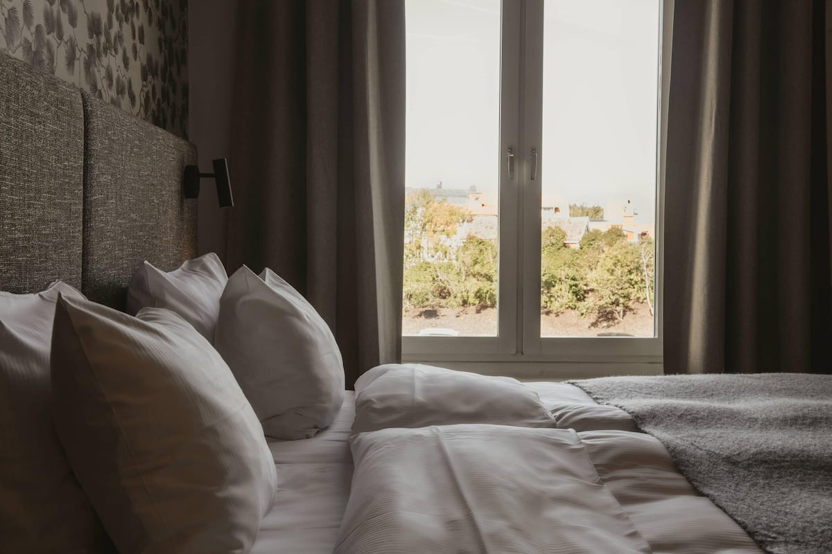Ett hotellrum med en säng, vita sängkläder, utsikt över omgivningen genom ett fönster och en sänggavel med ett mönstrat utseende.