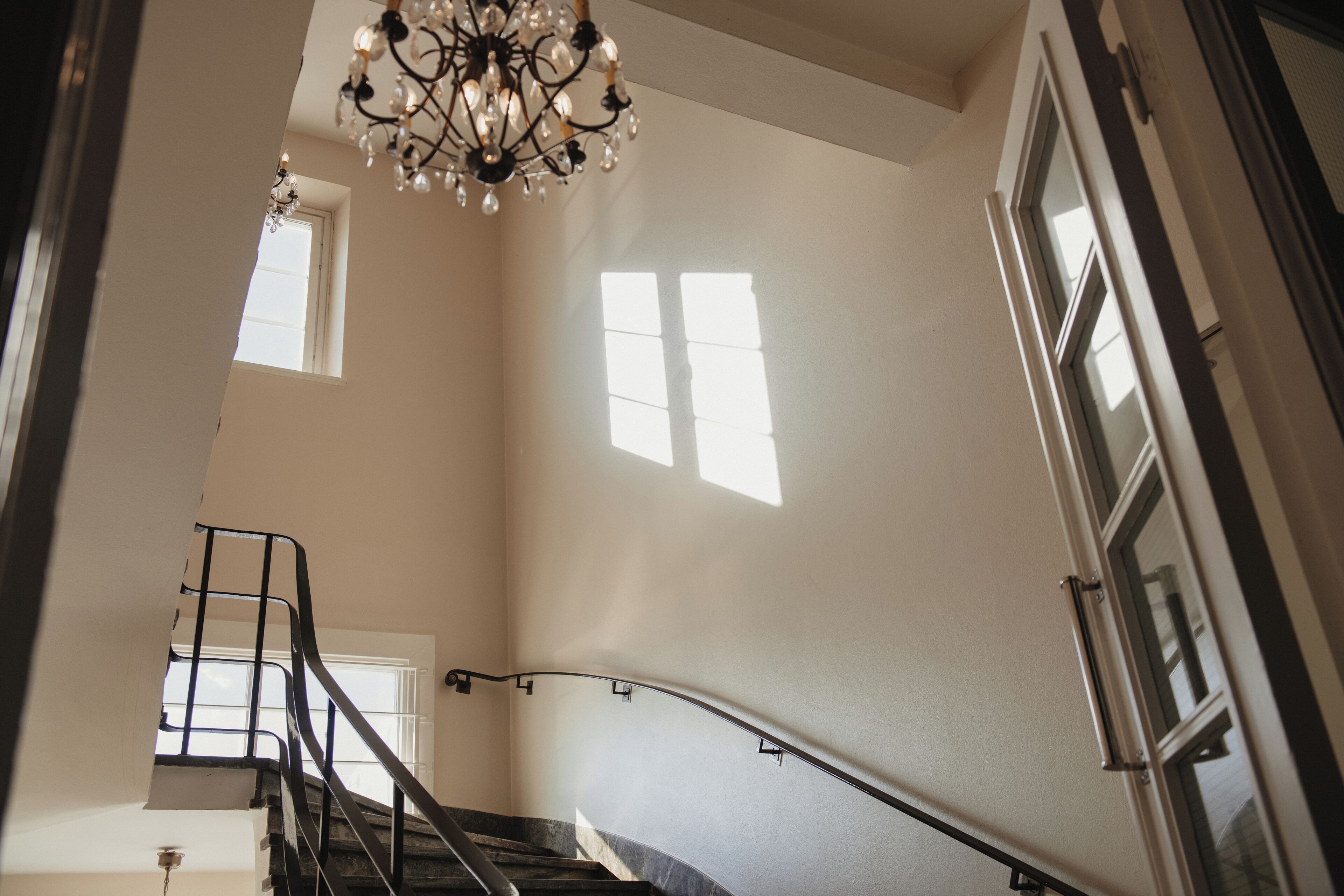 Una escalera con barandilla metálica, un candelabro y ventanas que dejan entrar luz natural.