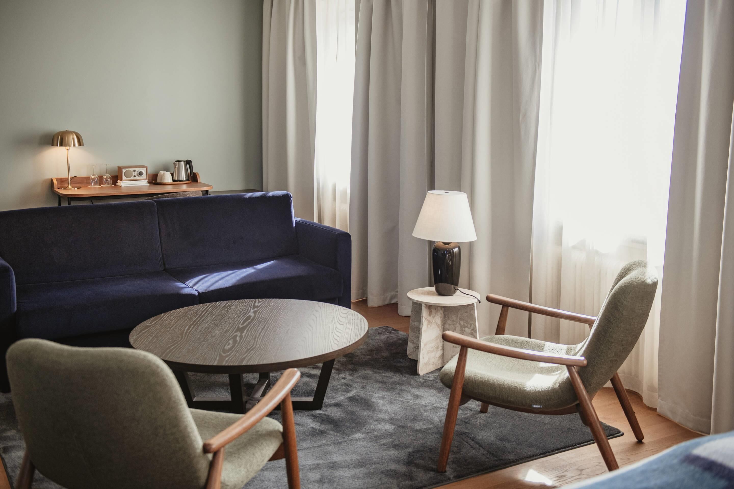 Una sala de estar moderna con un sofá azul oscuro, una mesa redonda de madera, dos sillones, una mesita auxiliar con una lámpara y una radio.