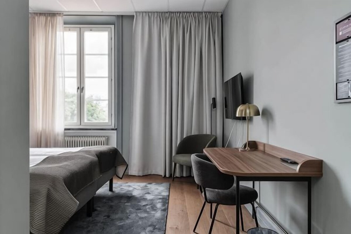 Ett hotellrum med en säng, ett skrivbord, en stol, en tv och ett fönster med gardiner.