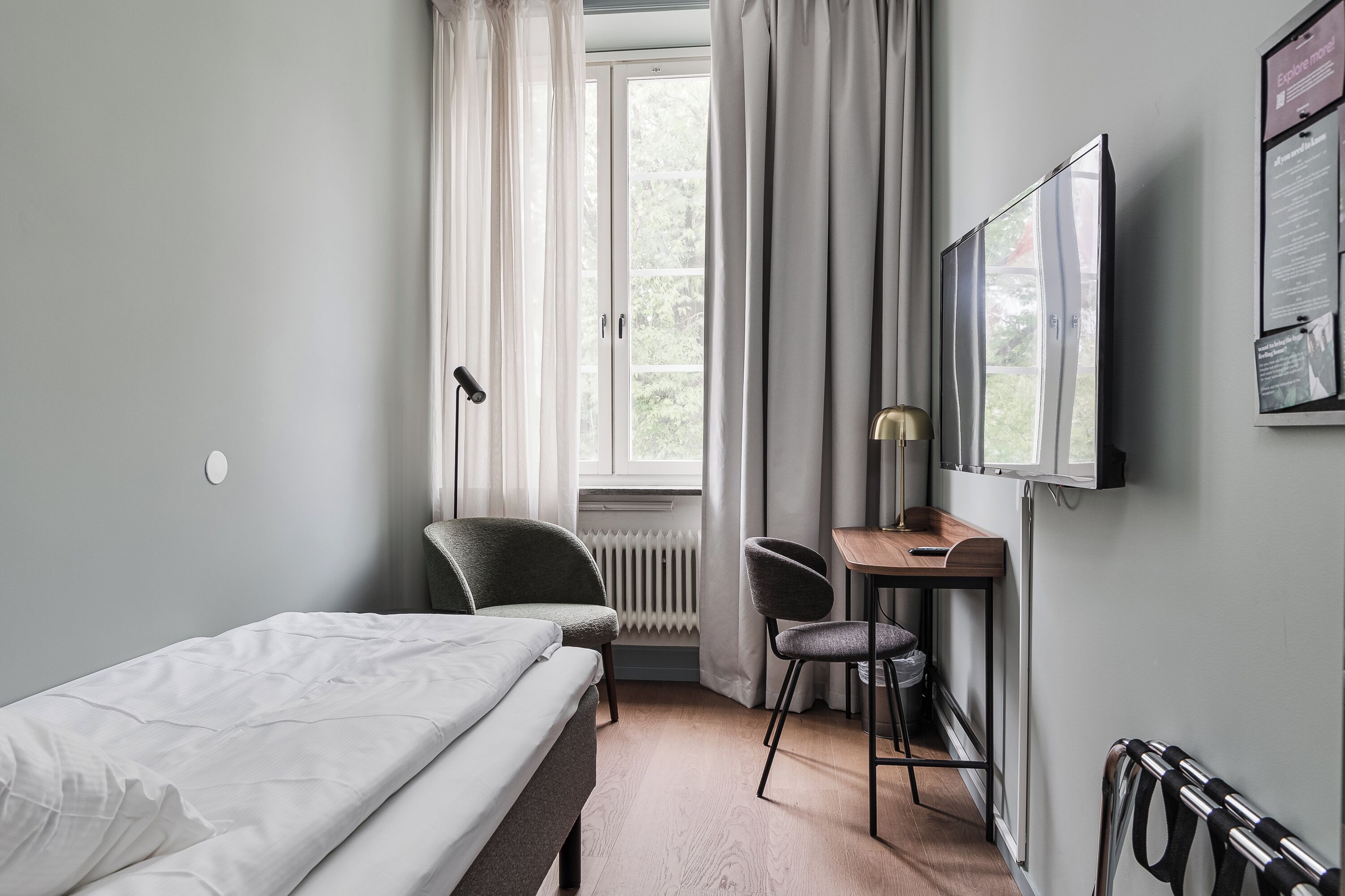 A hotel room with a bed, a desk, two chairs, a radiator, a window with curtains, and a mounted television.