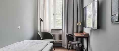 A hotel room with a bed, a desk, two chairs, a radiator, a window with curtains, and a mounted television.