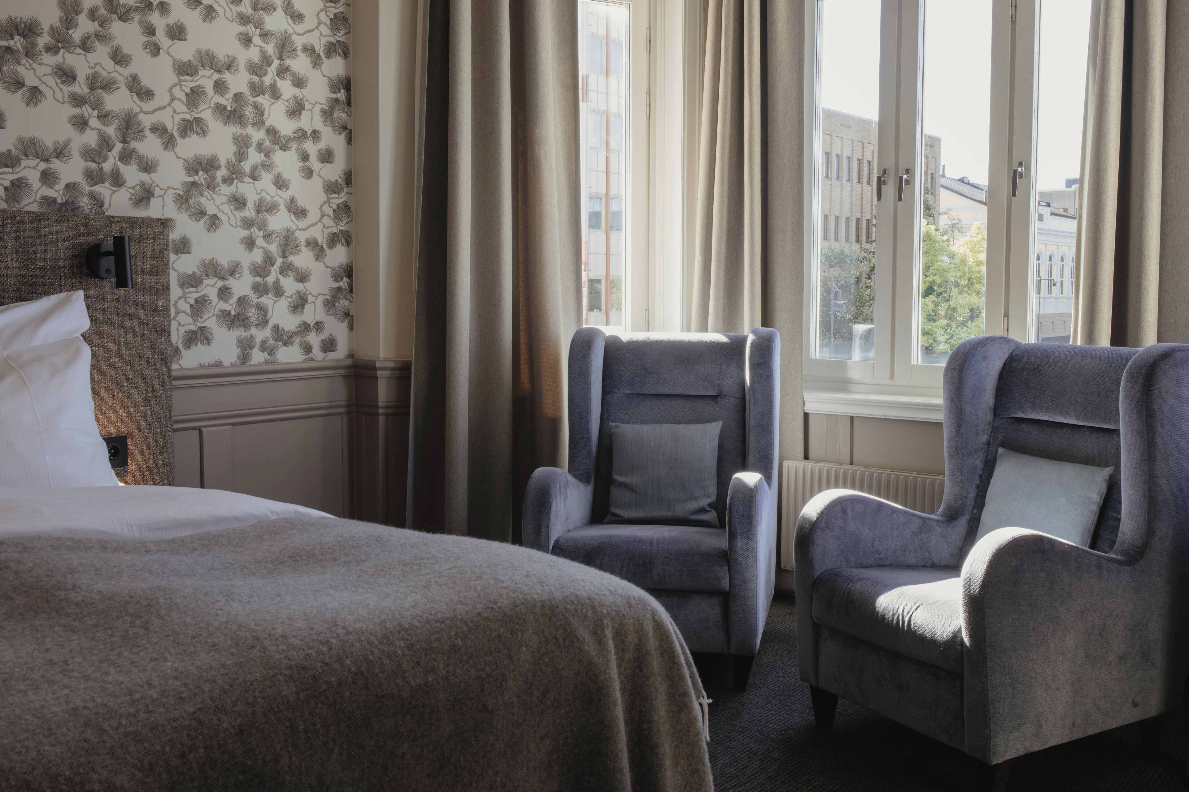 Une chambre d’hôtel avec un lit, deux fauteuils, une fenêtre avec des rideaux et un papier peint à motifs floraux.