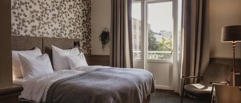 Habitación de hotel con una cama grande, una silla, una lámpara y vistas de la ciudad a través de una ventana.