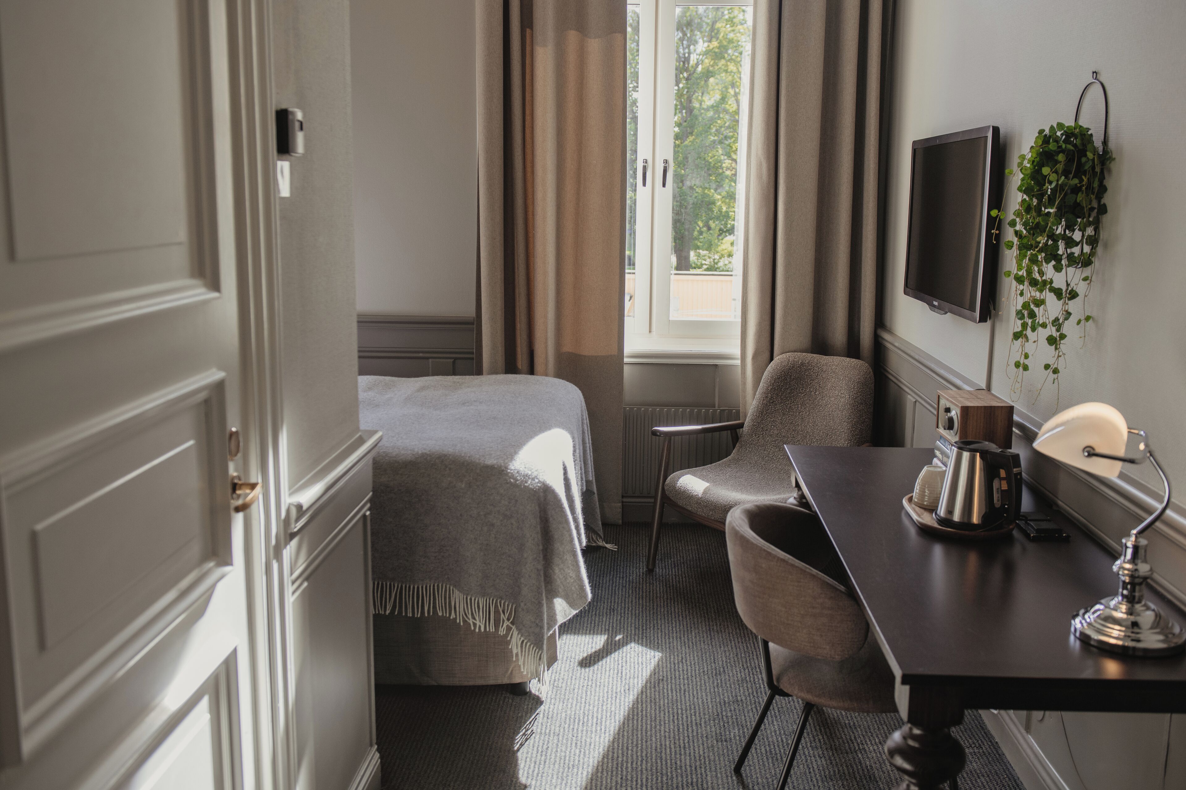 A hotel room with a bed, a desk with a lamp, a chair, a television, and a window with curtains.