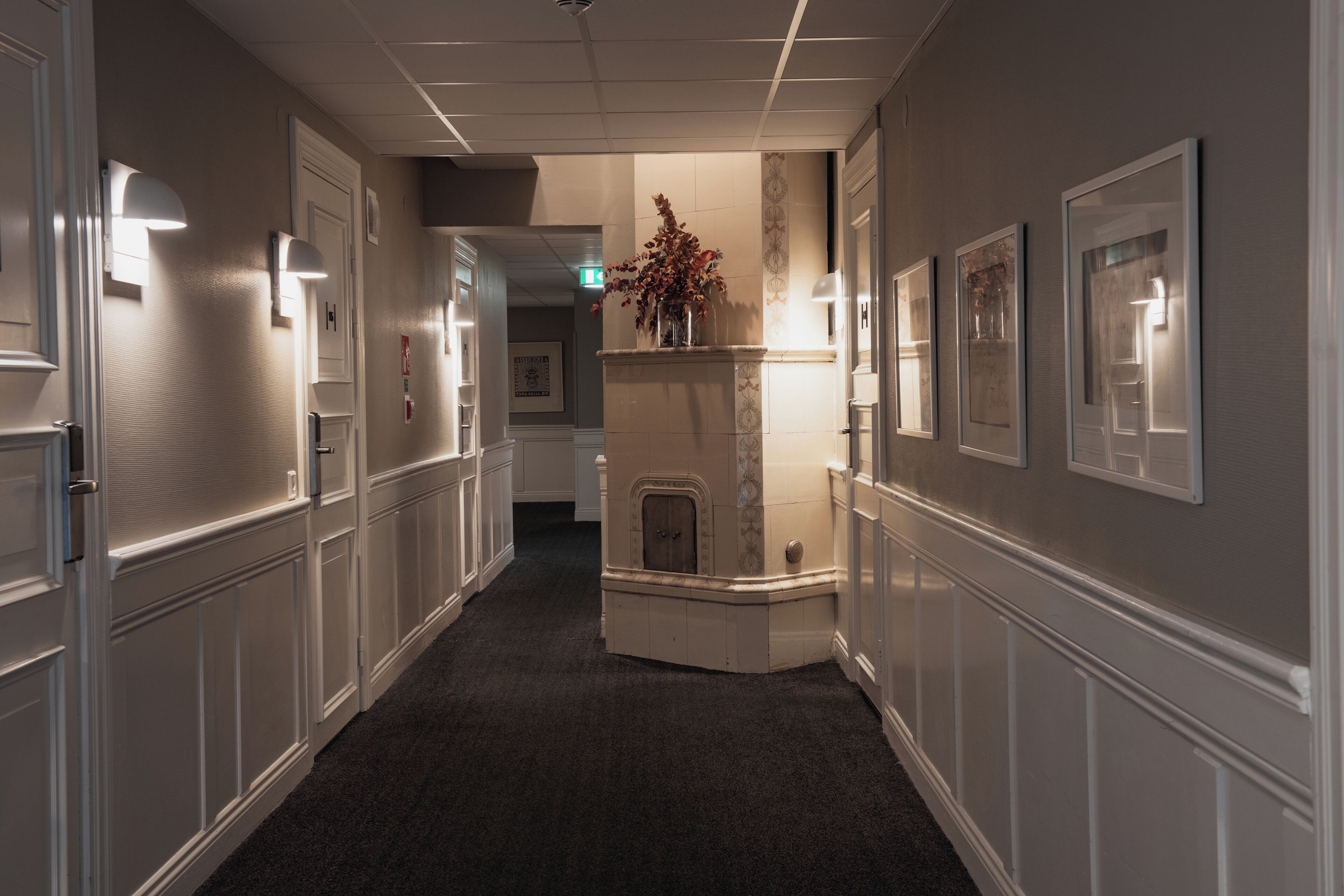 A hallway with white paneling, framed artwork on the walls, and a fireplace with a vase of flowers.
