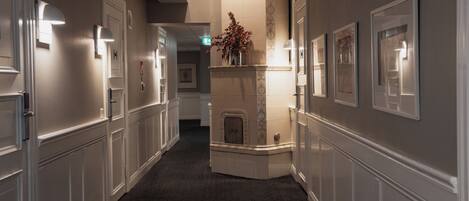 A hallway with white paneling, framed artwork on the walls, and a fireplace with a vase of flowers.
