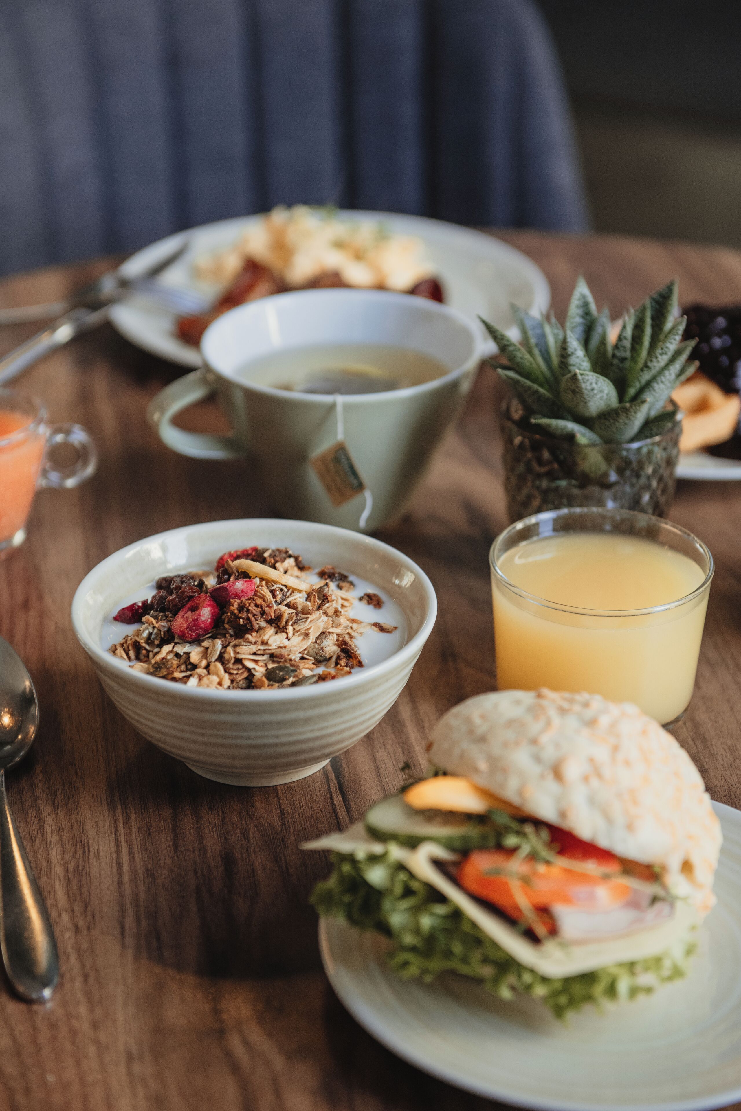 Desayuno con un tazón de granola, un vaso de jugo de naranja, un sándwich y una taza de té sobre una mesa de madera.