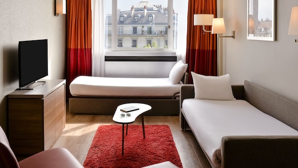 Une chambre d’hôtel avec un lit, un canapé, une télévision, une petite table et une fenêtre avec des rideaux.
