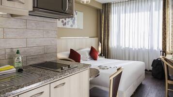 A hotel room with a kitchenette, a bed with red pillows, a desk, and a view of the outdoors.