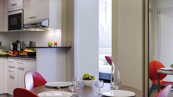 Une cuisine moderne équipée d’un four à micro-ondes, d’une cuisinière et d’un panier de fruits posé sur le comptoir. On y trouve également un coin repas avec une table dressée pour deux personnes, des chaises rouges et une vue sur un balcon.