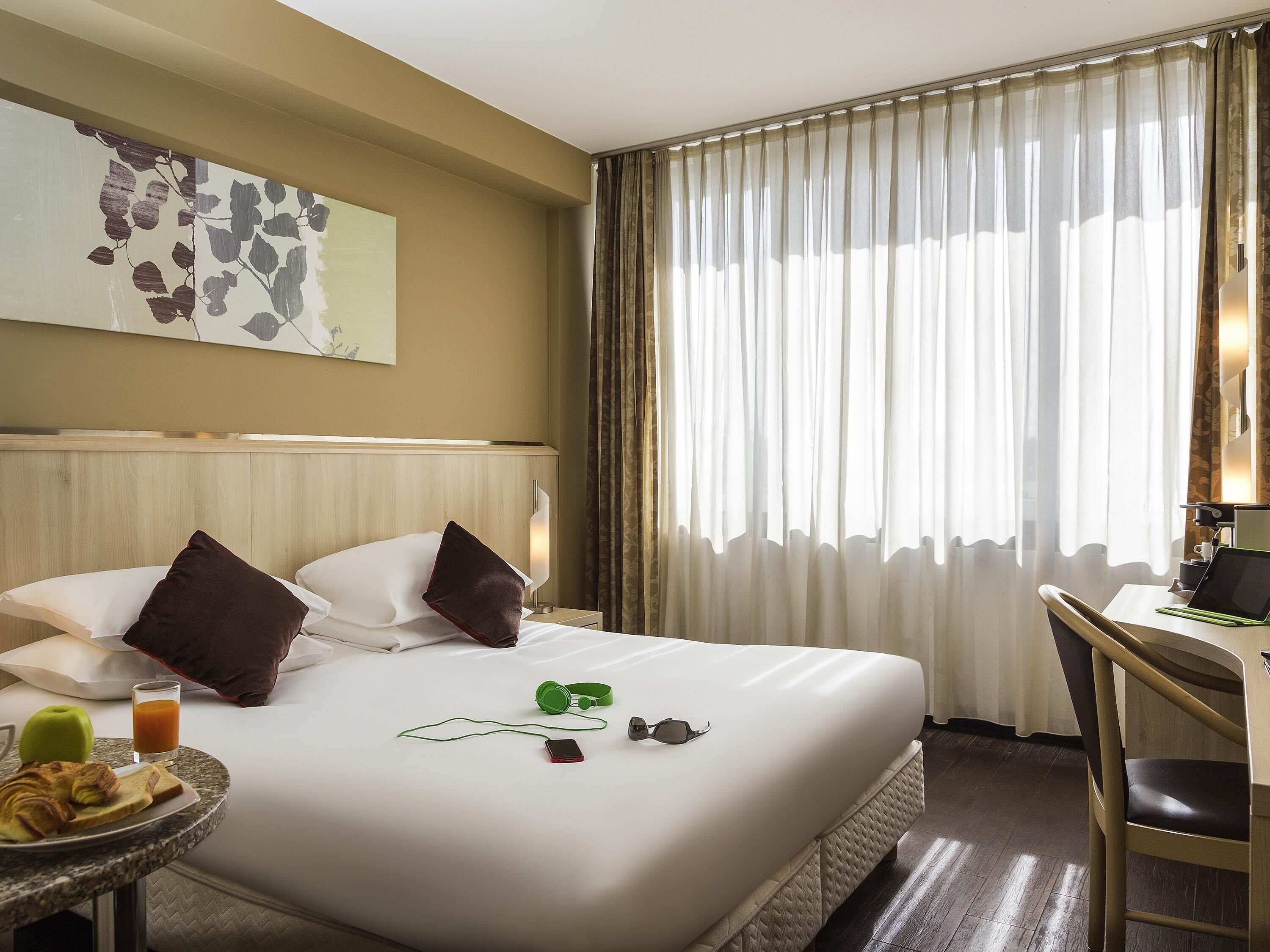 A hotel room with a bed, a desk, a chair, a small table with breakfast, and a window with curtains.