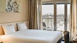 Une chambre d’hôtel avec un grand lit, une vue sur la ville depuis la fenêtre et un balcon avec des chaises.