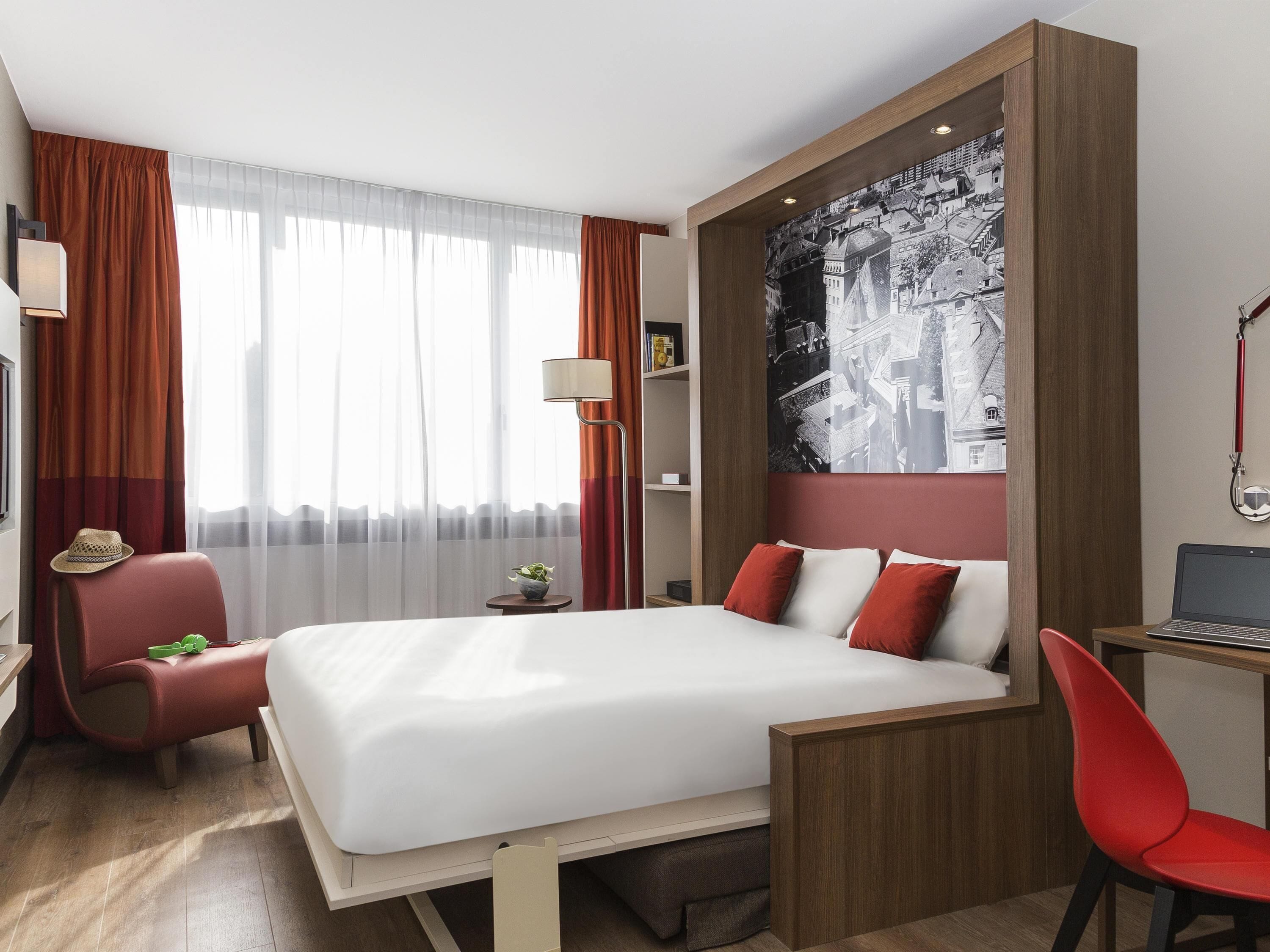 Ein modernes Hotelzimmer mit einem großen Bett, einem Schreibtisch, einem Stuhl, einer Lampe und einem Stadtbild an der Wand.