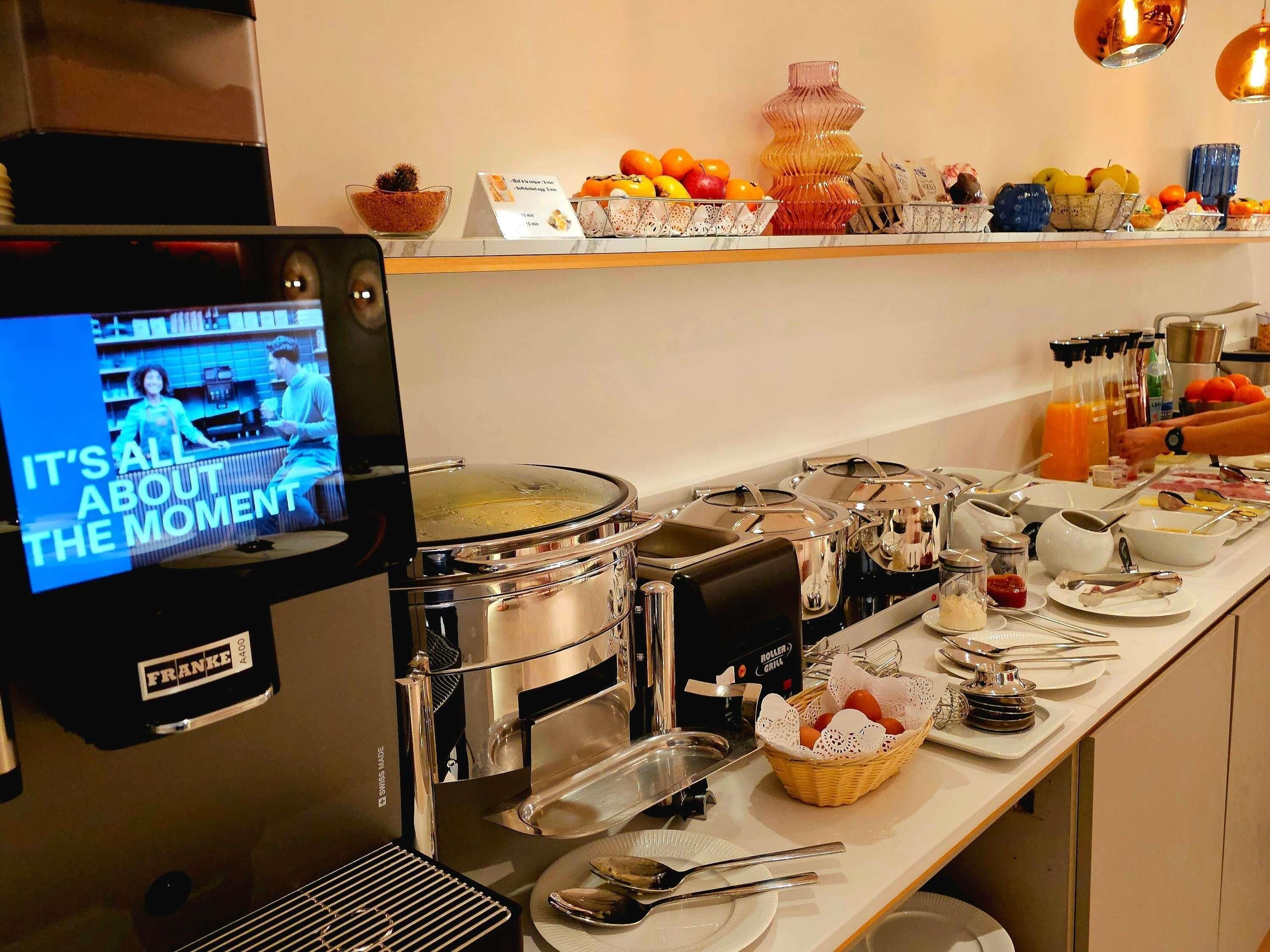 Een buffetopstelling met diverse gerechten, een koffieapparaat en een televisiescherm waarop een kookprogramma draait.