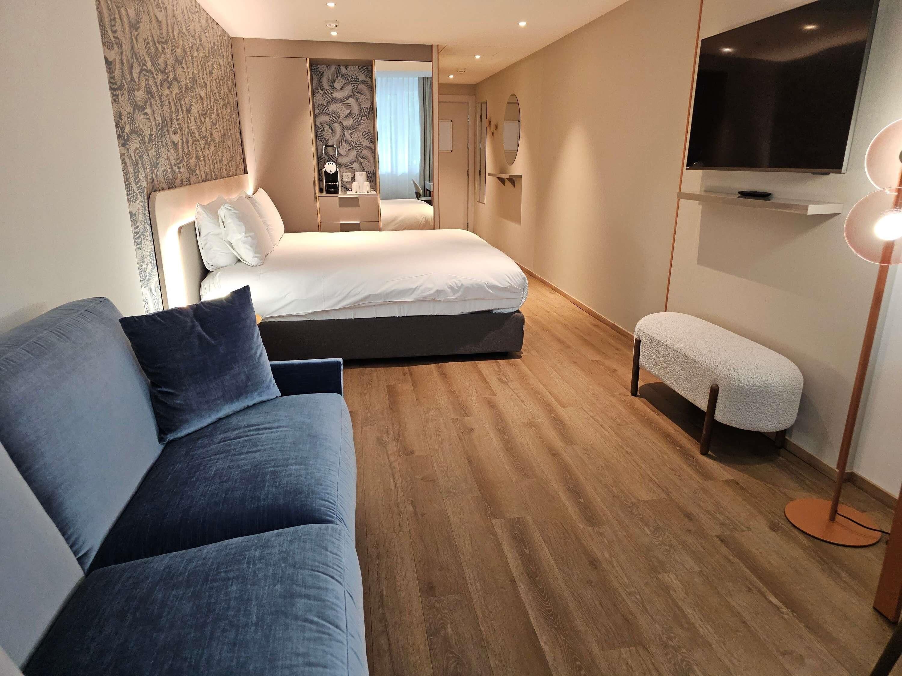 A modern hotel room with a bed, a sofa, a bench, a TV, and a mirror.