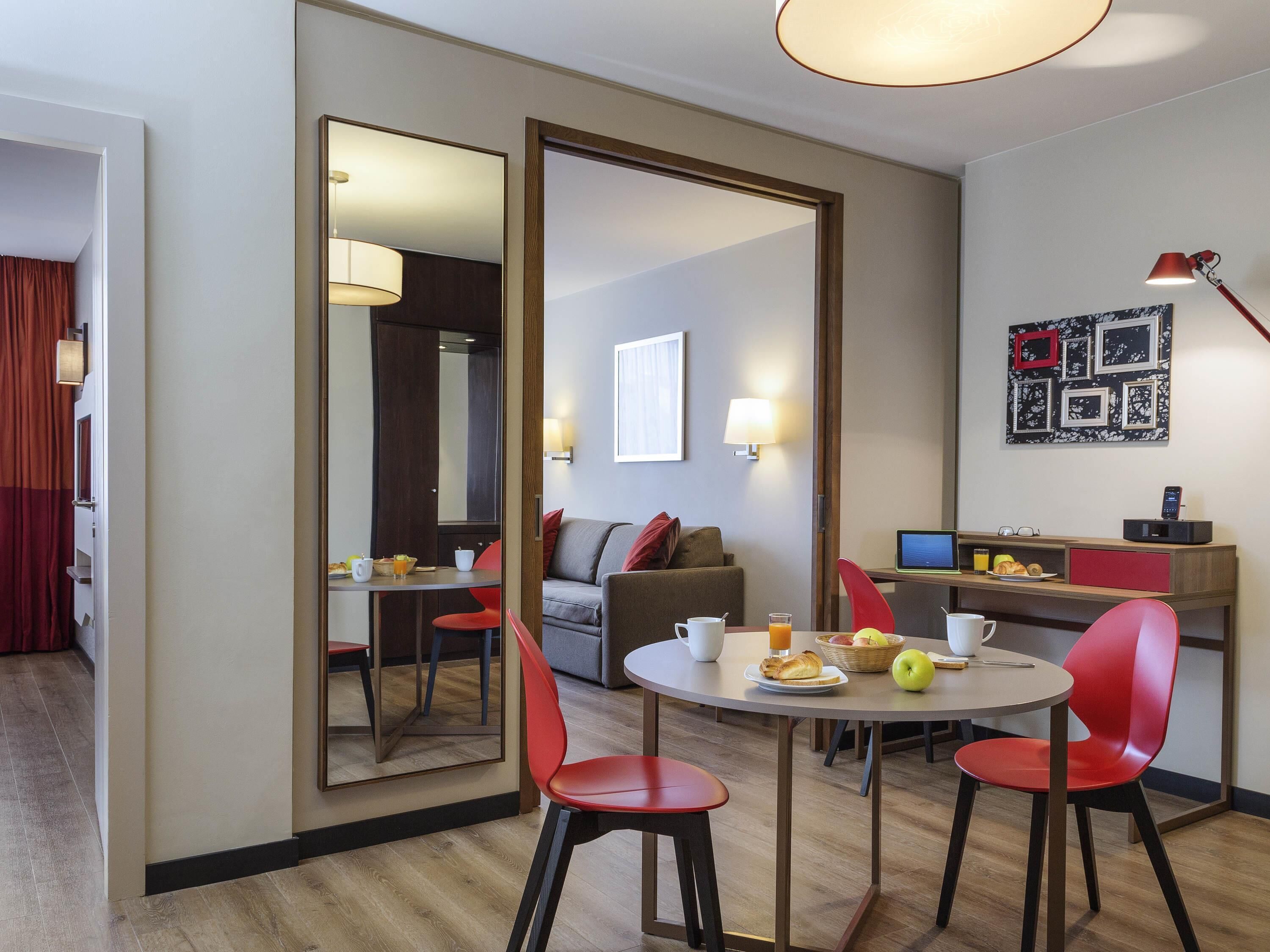 Una habitación de hotel con comedor, sofá, escritorio con computadora y un espejo grande.