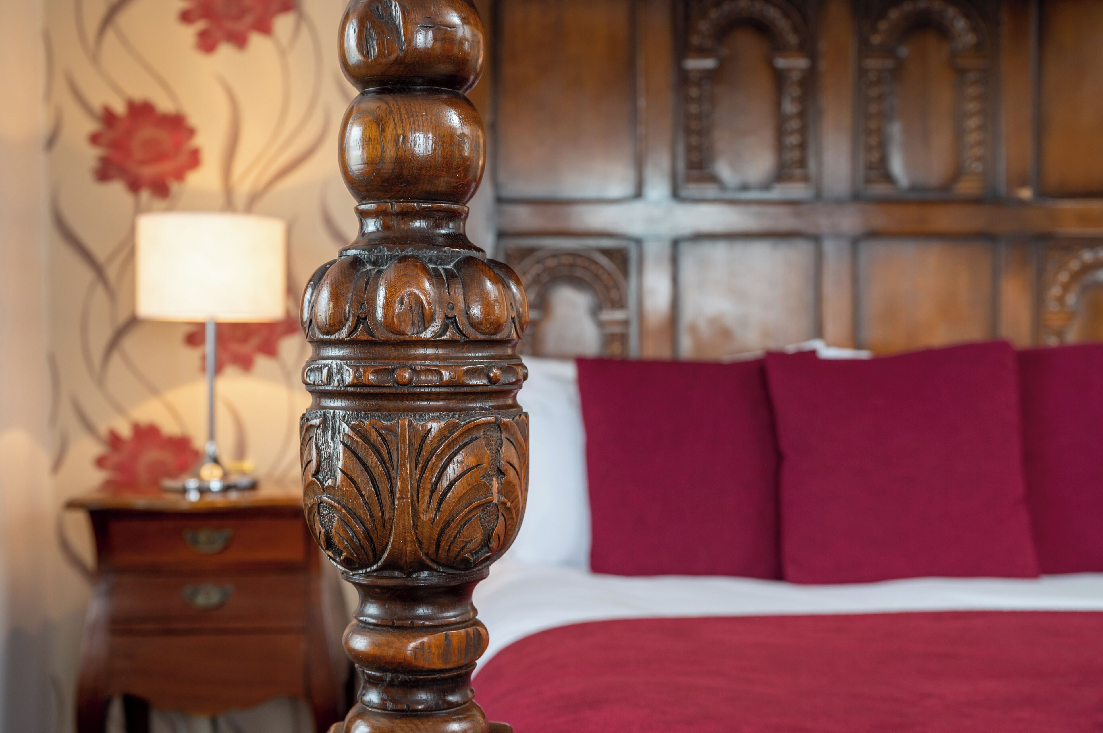 A wooden bed frame with intricate carvings, a red bedspread, and a nightstand with a lamp.
